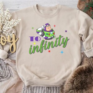 May include: A beige sweatshirt featuring a cartoon image of Buzz Lightyear saluting, with the words "To infinity" in green script. Colorful stars and accents surround the graphic. The sweatshirt is a casual, comfortable style.