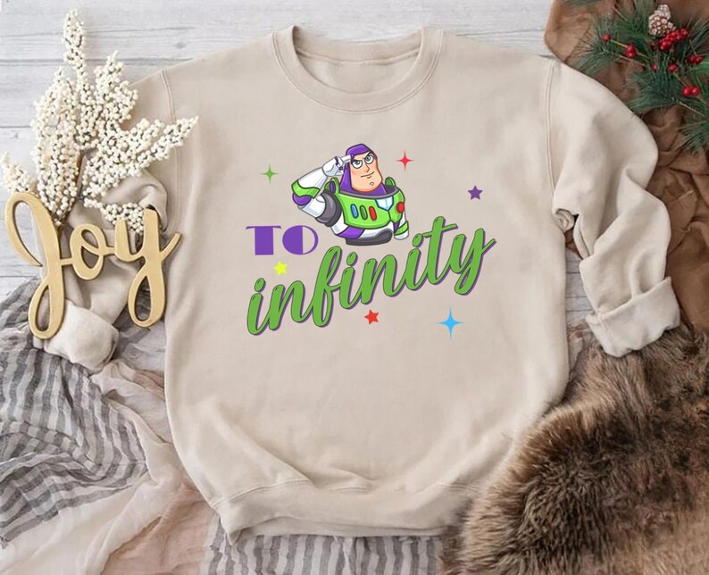 May include: A beige sweatshirt featuring a cartoon image of Buzz Lightyear saluting, with the words "To infinity" in green script. Colorful stars and accents surround the graphic. The sweatshirt is a casual, comfortable style.