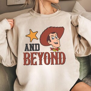 May include: A beige sweatshirt featuring a cartoon image of a cowboy with a sheriff's badge and the words "AND BEYOND" in a western-style font. The cowboy is wearing a brown hat and a red bandana.