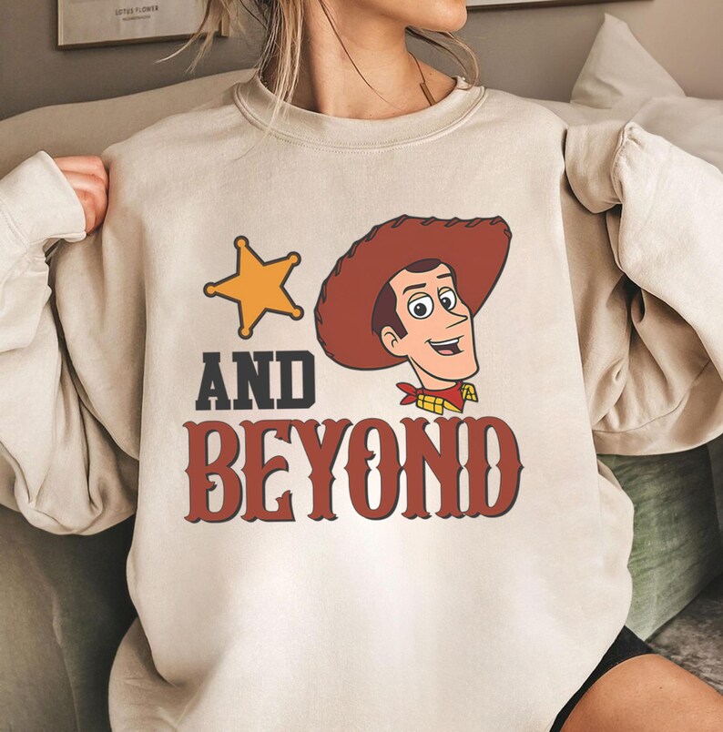 May include: A beige sweatshirt featuring a cartoon image of a cowboy with a sheriff's badge and the words "AND BEYOND" in a western-style font. The cowboy is wearing a brown hat and a red bandana.