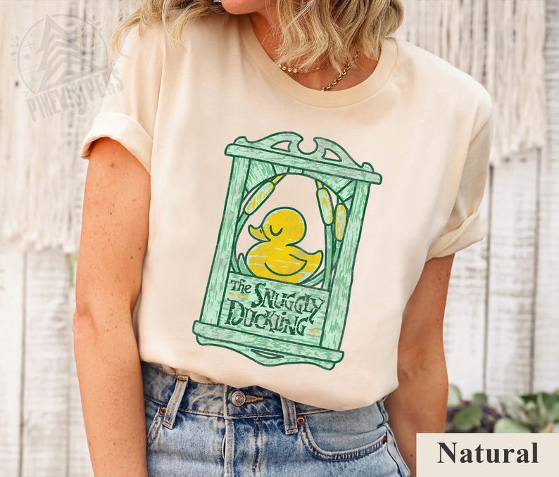 May include: Cream-colored t-shirt featuring a green framed design with a yellow duck illustration and the text "The Snuggly Duckling." The design includes cattails. The shirt is made of a soft material and has rolled-up sleeves.