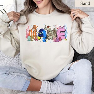 May include: Sand-colored sweatshirt with the word "NAME" in colorful, whimsical lettering. The letters are decorated with sea creatures and coral, creating a playful, underwater theme. The sweatshirt is a neutral color, making the design stand out.