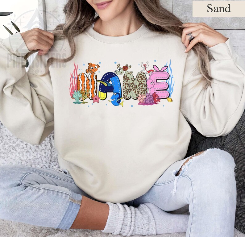 May include: Sand-colored sweatshirt with the word "NAME" in colorful, whimsical lettering. The letters are decorated with sea creatures and coral, creating a playful, underwater theme. The sweatshirt is a neutral color, making the design stand out.