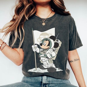 May include: A dark gray t-shirt with a graphic of Mickey Mouse dressed as an astronaut planting a flag on the moon. The flag has a circle with the letters "BB" inside.