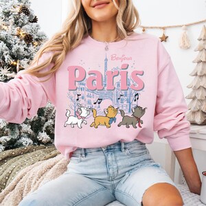 Puede incluir: Una sudadera rosa claro con la palabra "Par&iacute;s" en letras grandes, la Torre Eiffel y notas musicales. Debajo del texto hay tres gatitos de dibujos animados. La palabra "Bonjour" est&aacute; encima de la palabra "Par&iacute;s".