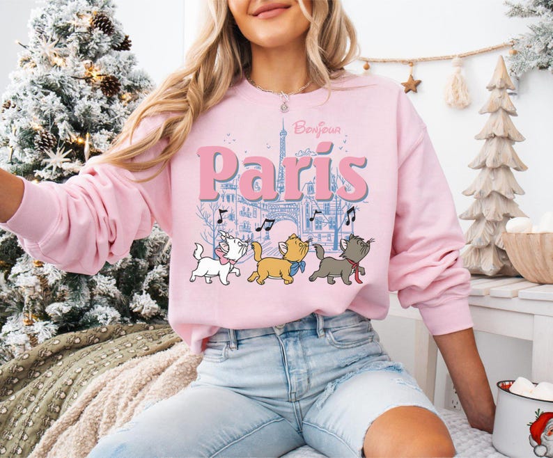 Puede incluir: Una sudadera rosa claro con la palabra "Par&iacute;s" en letras grandes, la Torre Eiffel y notas musicales. Debajo del texto hay tres gatitos de dibujos animados. La palabra "Bonjour" est&aacute; encima de la palabra "Par&iacute;s".