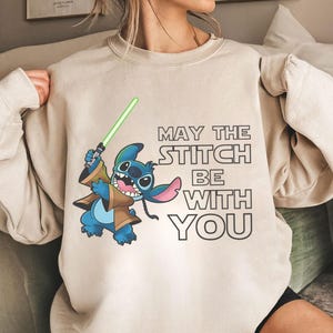 May include: Beige sweatshirt featuring a cartoon Stitch character holding a green lightsaber. The design includes the text "MAY THE STITCH BE WITH YOU" in black. The sweatshirt is a neutral color.