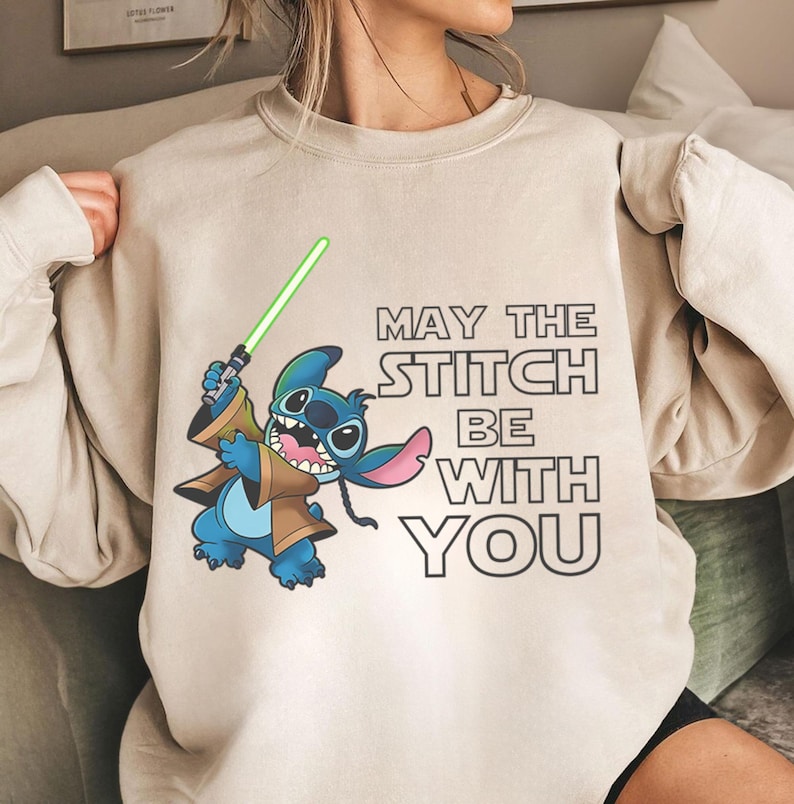 May include: Beige sweatshirt featuring a cartoon Stitch character holding a green lightsaber. The design includes the text "MAY THE STITCH BE WITH YOU" in black. The sweatshirt is a neutral color.