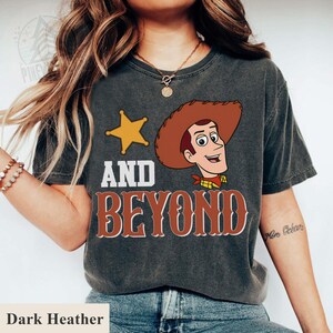 May include: Dark heather t-shirt featuring a cartoon image of Woody from Toy Story, a gold star, and the words "AND BEYOND" in a western-style font. The shirt is a dark gray color.