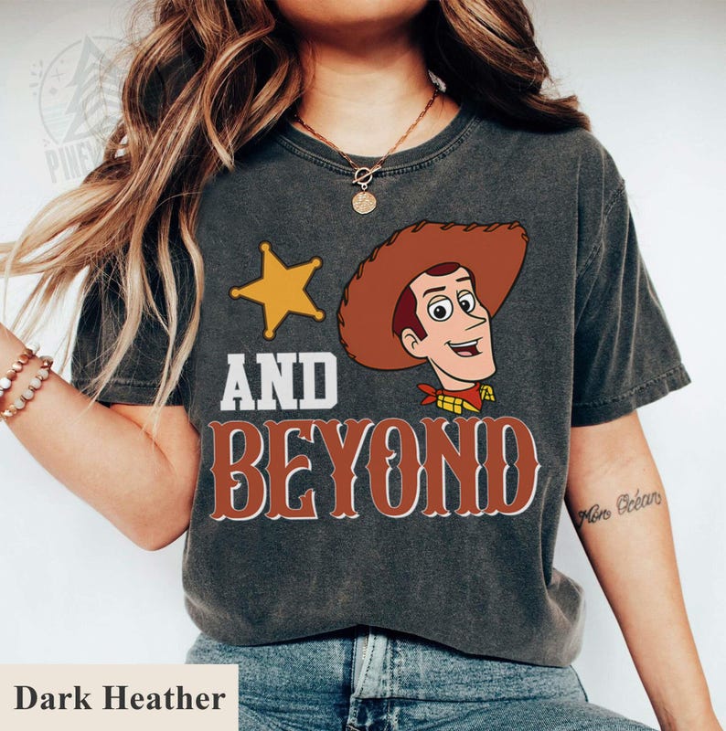 May include: Dark heather t-shirt featuring a cartoon image of Woody from Toy Story, a gold star, and the words "AND BEYOND" in a western-style font. The shirt is a dark gray color.