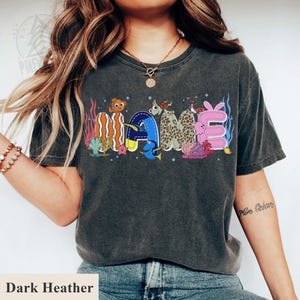 May include: Dark heather gray t-shirt with the word "NAME" in colorful, cartoonish, underwater-themed letters. The letters are decorated with sea creatures and coral. The shirt is a comfortable, casual style.