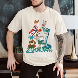 Puede incluir: Camiseta color crema con un gr&aacute;fico colorido de personajes de la pel&iacute;cula animada "Zootopia". El dise&ntilde;o incluye un zorro, un conejo, una serpiente y otros personajes, con el t&iacute;tulo de la pel&iacute;cula en azul y verde. La camiseta tambi&eacute;n tiene el texto "Hello Clark."