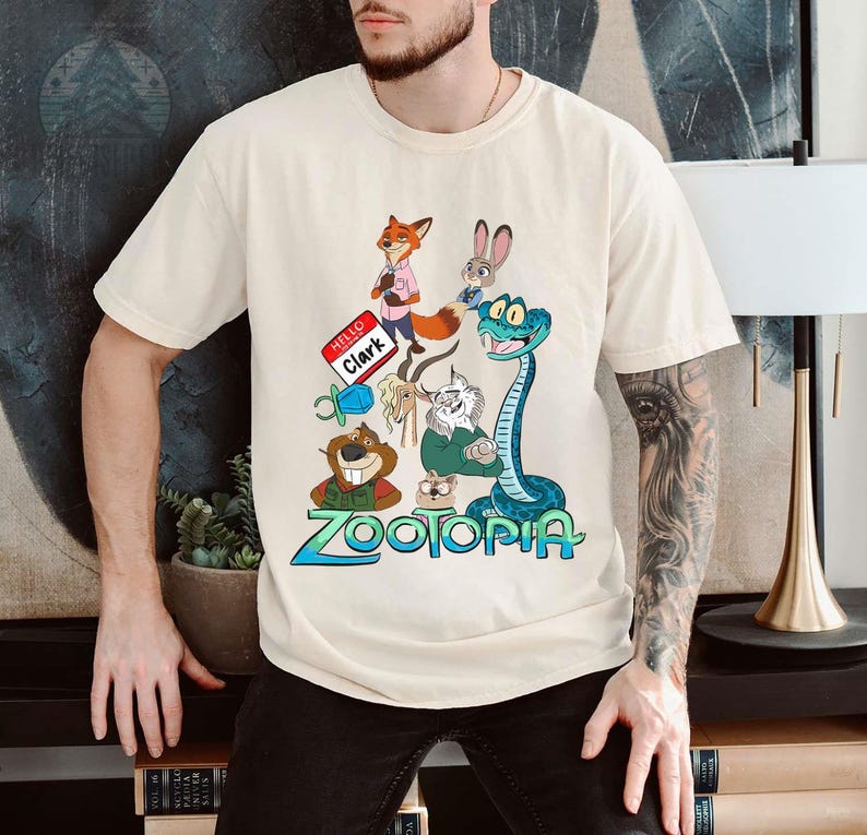 Puede incluir: Camiseta color crema con un gr&aacute;fico colorido de personajes de la pel&iacute;cula animada "Zootopia". El dise&ntilde;o incluye un zorro, un conejo, una serpiente y otros personajes, con el t&iacute;tulo de la pel&iacute;cula en azul y verde. La camiseta tambi&eacute;n tiene el texto "Hello Clark."