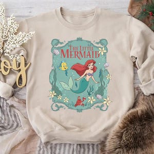 May include: Beige crewneck sweatshirt featuring a colorful graphic of Ariel from The Little Mermaid. The graphic includes Ariel, Flounder, Sebastian, and the words "The Little Mermaid" in a decorative frame. The sweatshirt is made of a soft material.