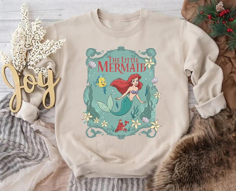 May include: Beige crewneck sweatshirt featuring a colorful graphic of Ariel from The Little Mermaid. The graphic includes Ariel, Flounder, Sebastian, and the words "The Little Mermaid" in a decorative frame. The sweatshirt is made of a soft material.