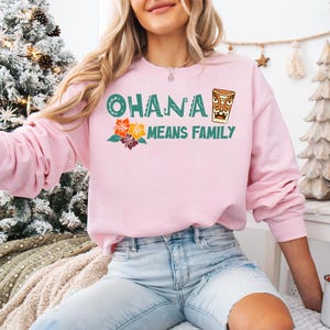 May include: A light pink sweatshirt with the text "OHANA MEANS FAMILY" in green and orange. The design includes a tiki mug and tropical flowers. The sweatshirt is long-sleeved and made of a soft material.