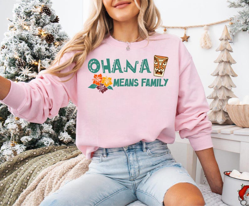 May include: A light pink sweatshirt with the text "OHANA MEANS FAMILY" in green and orange. The design includes a tiki mug and tropical flowers. The sweatshirt is long-sleeved and made of a soft material.