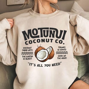 May include: Beige sweatshirt with the text "MOTUNUI COCONUT CO." in black. The design includes illustrations of coconuts and text about coconut products. The phrase "IT'S ALL YOU NEED" is at the bottom.