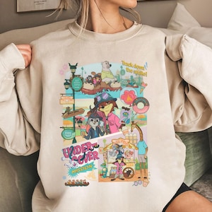 May include: Beige sweatshirt featuring a colorful cartoon graphic with the text "Undercover Mission" and "Track down the snake!". The graphic includes various animal characters and amusement park elements.