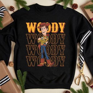 Puede incluir: Sudadera negra con el personaje Woody de Toy Story. El dise&ntilde;o presenta la palabra "WOODY" apilada en un estilo retro, con una ilustraci&oacute;n de Woody con su atuendo de vaquero. Decoraciones navide&ntilde;as rodean la sudadera.