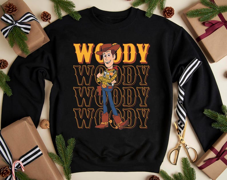Puede incluir: Sudadera negra con el personaje Woody de Toy Story. El dise&ntilde;o presenta la palabra "WOODY" apilada en un estilo retro, con una ilustraci&oacute;n de Woody con su atuendo de vaquero. Decoraciones navide&ntilde;as rodean la sudadera.