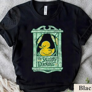 May include: Black t-shirt featuring a yellow duck graphic inside a green frame with the text "The Snuggly Duckling". The design includes cattails and a decorative border. The shirt is made of a soft material.