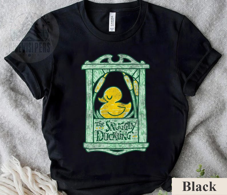 May include: Black t-shirt featuring a yellow duck graphic inside a green frame with the text "The Snuggly Duckling". The design includes cattails and a decorative border. The shirt is made of a soft material.