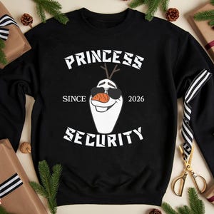 May include: Black sweatshirt with the text "PRINCESS SECURITY" and an illustration of Olaf wearing sunglasses. The text "SINCE 2026" is also included. The sweatshirt is surrounded by gift boxes, pine needles, and pine cones.