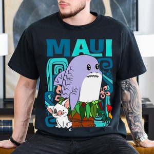 Puede incluir: Una camiseta negra con una ilustraci&oacute;n de dibujos animados de Maui de la pel&iacute;cula Moana. La ilustraci&oacute;n presenta a Maui como un tibur&oacute;n con un collar de flores y un cerdo y un gallo a su lado. El texto "MAUI" est&aacute; impreso en azul y verde sobre la ilustraci&oacute;n.