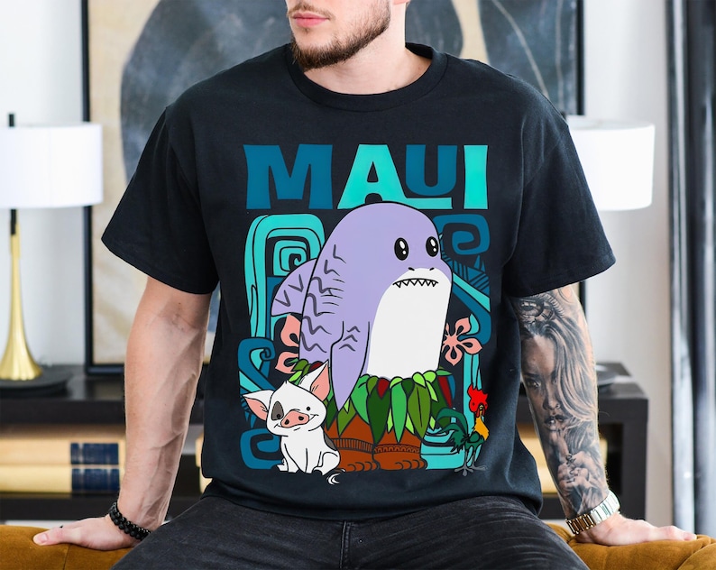 Puede incluir: Una camiseta negra con una ilustraci&oacute;n de dibujos animados de Maui de la pel&iacute;cula Moana. La ilustraci&oacute;n presenta a Maui como un tibur&oacute;n con un collar de flores y un cerdo y un gallo a su lado. El texto "MAUI" est&aacute; impreso en azul y verde sobre la ilustraci&oacute;n.