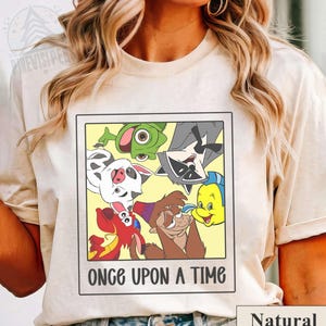 Op de afbeelding: Cr&egrave;mekleurig T-shirt met een ingelijste afbeelding van stripfiguren. De afbeelding bevat een kikker, een wasbeer, een varken, een vis en andere personages. De tekst "ONCE UPON A TIME" staat eronder. Het woord "Natural" staat rechtsonder.