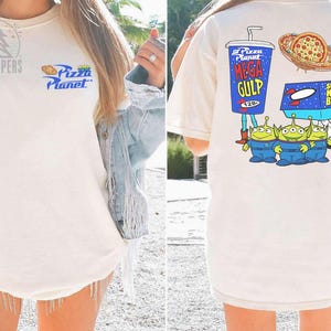 May include: Cream-colored t-shirt featuring the Pizza Planet logo on the front. The back of the shirt displays a Mega Gulp drink, pizza, a Super Nova Burger, and three green alien characters in blue uniforms.