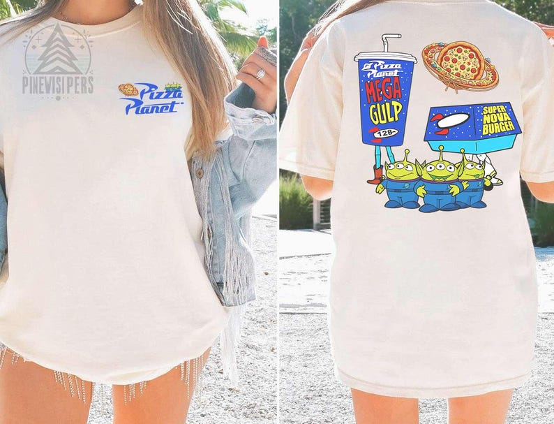 May include: Cream-colored t-shirt featuring the Pizza Planet logo on the front. The back of the shirt displays a Mega Gulp drink, pizza, a Super Nova Burger, and three green alien characters in blue uniforms.