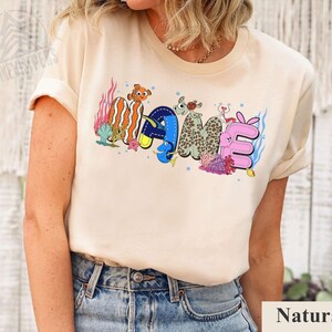May include: A beige t-shirt with the word "NAME" in colorful, cartoonish letters. The letters are designed with an ocean theme, featuring fish and coral. The shirt is made of a soft material and has short sleeves.