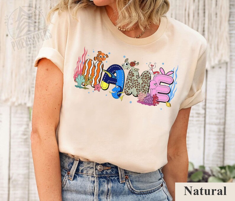 May include: A beige t-shirt with the word "NAME" in colorful, cartoonish letters. The letters are designed with an ocean theme, featuring fish and coral. The shirt is made of a soft material and has short sleeves.