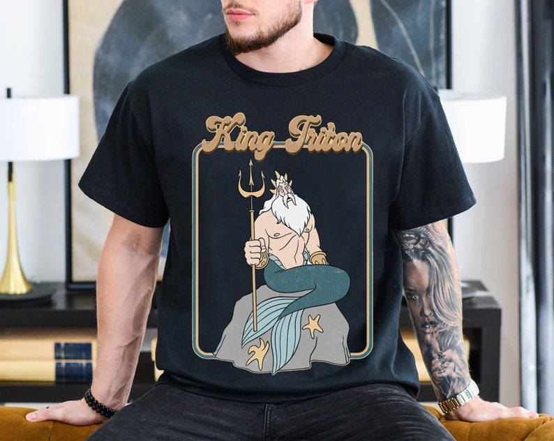 May include: A black t-shirt featuring a retro-style graphic of King Triton from Disney's The Little Mermaid. The graphic depicts King Triton sitting on a rock with his trident and a blue tail.
