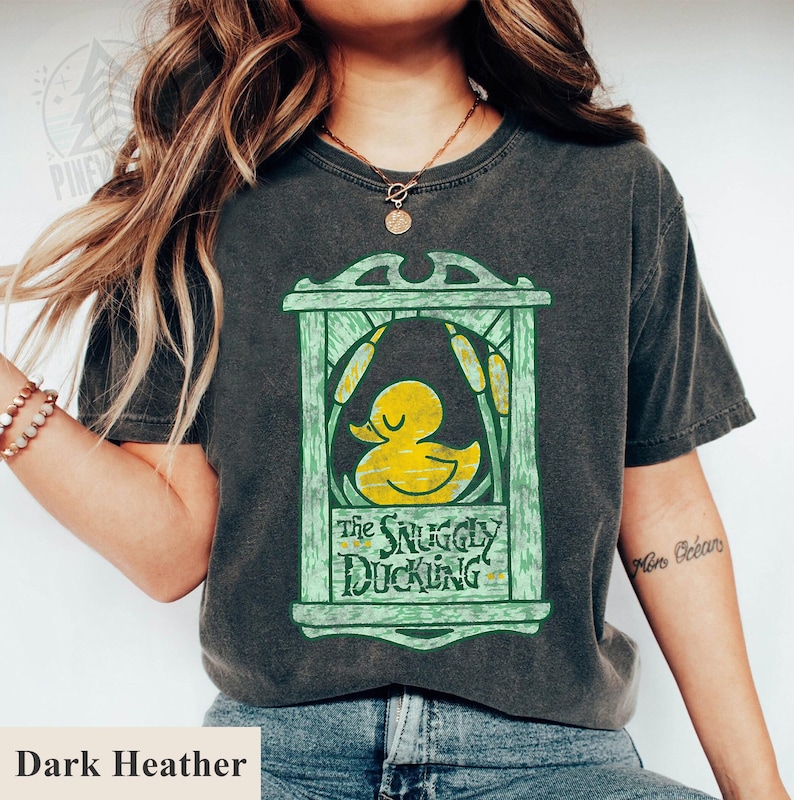 May include: Dark heather gray t-shirt featuring a yellow duck graphic inside a green frame with the text "The Snuggly Duckling". The design includes cattails and a vintage aesthetic.