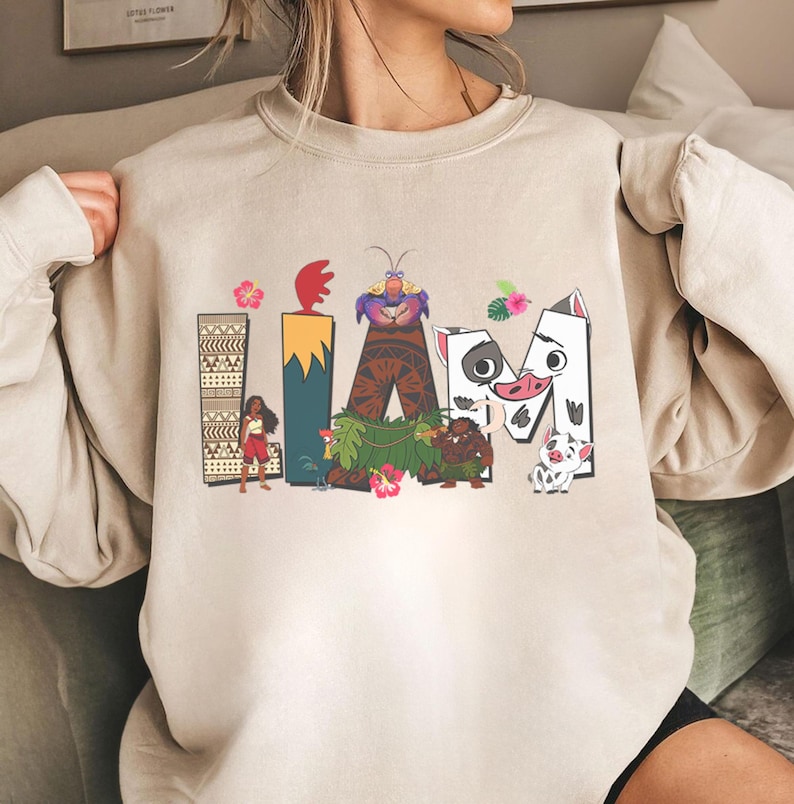 May include: Beige sweatshirt with the word "LIA M" in a colourful cartoon design. The letters are decorated with characters and elements from a popular animated film, including a woman, a crab, and a pig.