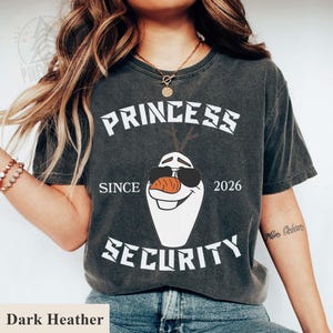 May include: Dark heather gray t-shirt featuring a graphic of Olaf from Frozen wearing sunglasses. The text reads "PRINCESS SECURITY SINCE 2026". A fun and playful design for fans of the movie.