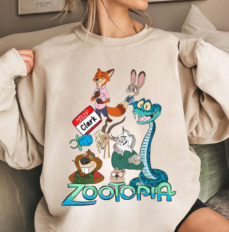 Puede incluir: Sudadera beige de cuello redondo con un gr&aacute;fico colorido de personajes de la pel&iacute;cula animada "Zootopia". El dise&ntilde;o incluye un zorro, un conejo, una serpiente y otros animales, con la palabra "Zootopia" en verde y azul. Una etiqueta con el nombre dice "Hello Clark."