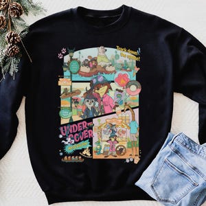 May include: Black sweatshirt featuring a colorful cartoon graphic with the text "Undercover Mission" and "Track down the snake!". The graphic includes various animal characters in a theme park setting. A pair of blue jeans is folded next to the sweatshirt.