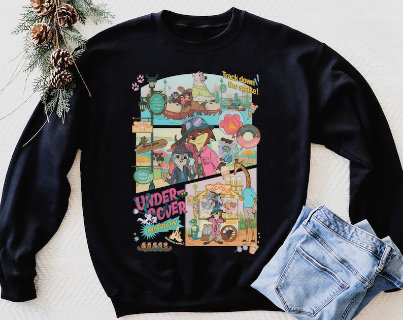 May include: Black sweatshirt featuring a colorful cartoon graphic with the text "Undercover Mission" and "Track down the snake!". The graphic includes various animal characters in a theme park setting. A pair of blue jeans is folded next to the sweatshirt.