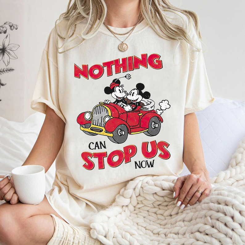 May include: A cream colored t-shirt with a graphic of Mickey and Minnie Mouse driving a red car. The text "NOTHING CAN STOP US NOW" is printed on the shirt.