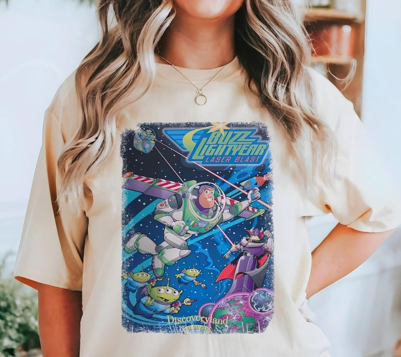 Puede incluir: Una camiseta de color beige claro con un gr&aacute;fico de estilo vintage de Buzz Lightyear de la franquicia Toy Story. El gr&aacute;fico presenta a Buzz Lightyear en su traje de ranger espacial, volando por el espacio con un disparo l&aacute;ser. El texto "Buzz Lightyear Laser Blast" aparece sobre el gr&aacute;fico, y "Discoveryland" aparece debajo del gr&aacute;fico.