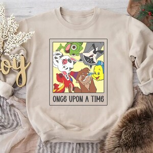 Op de afbeelding: Beige sweatshirt met een cartoonafbeelding van animatiepersonages in een gele lijst. De tekst "ONCE UPON A TIME" staat eronder gedrukt. De sweatshirt is gemaakt van zacht materiaal.