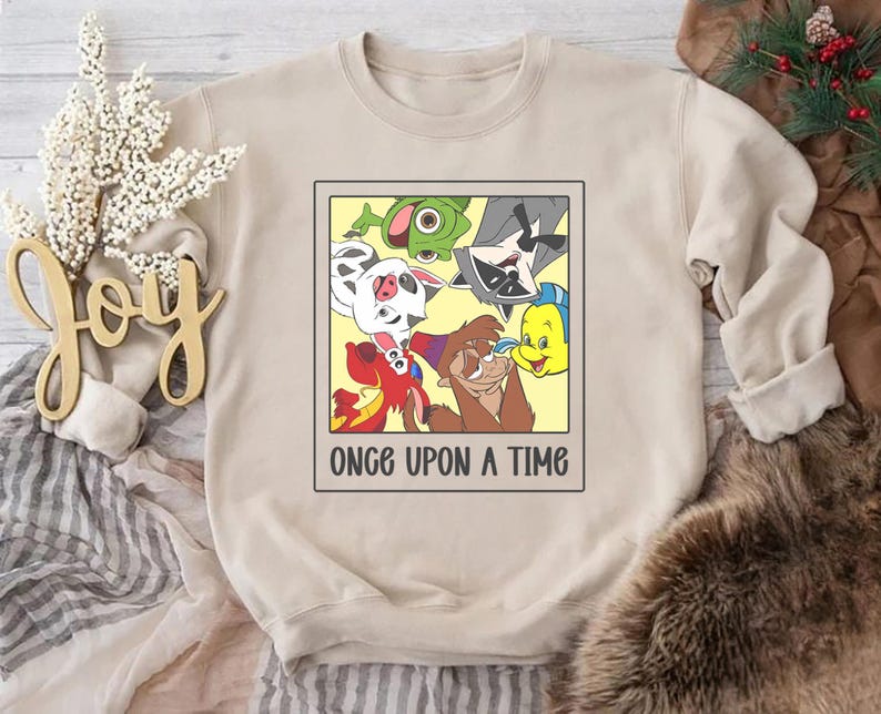 Op de afbeelding: Beige sweatshirt met een cartoonafbeelding van animatiepersonages in een gele lijst. De tekst "ONCE UPON A TIME" staat eronder gedrukt. De sweatshirt is gemaakt van zacht materiaal.