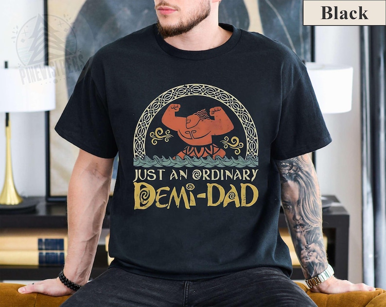 Op de afbeelding: Zwart T-shirt met een vintage-stijl illustratie van een gespierd figuur met een kroon en de tekst "Just an Ordinary Demi-Dad".