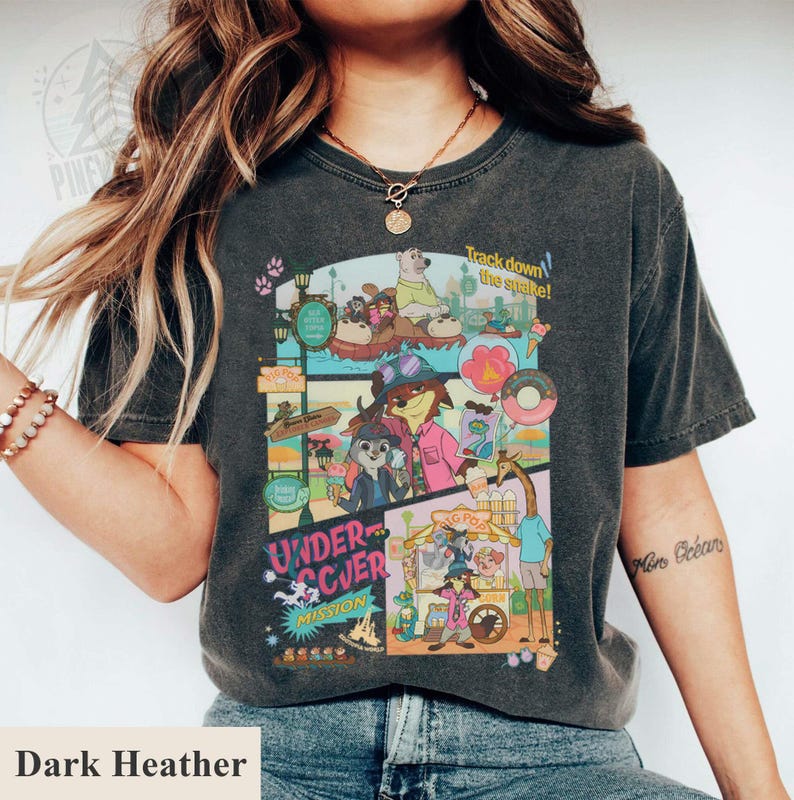 May include: Dark heather gray t-shirt featuring a colorful cartoon graphic with the text "Undercover Mission" and "Track down the snake!". The graphic includes various animal characters and food items. The shirt is a comfortable, casual style.