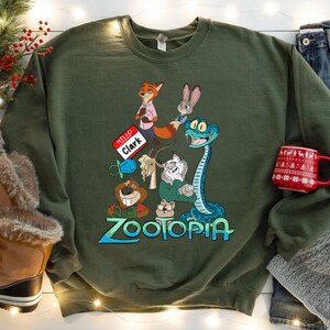 Puede incluir: Una sudadera verde oliva con personajes de la pel&iacute;cula animada Zootopia. El dise&ntilde;o incluye a Nick Wilde, Judy Hopps y otros personajes. La palabra "Zootopia" est&aacute; escrita en azul y verde. Una taza roja con un patr&oacute;n blanco est&aacute; a la derecha.