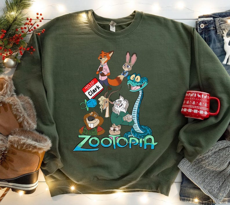 Puede incluir: Una sudadera verde oliva con personajes de la pel&iacute;cula animada Zootopia. El dise&ntilde;o incluye a Nick Wilde, Judy Hopps y otros personajes. La palabra "Zootopia" est&aacute; escrita en azul y verde. Una taza roja con un patr&oacute;n blanco est&aacute; a la derecha.